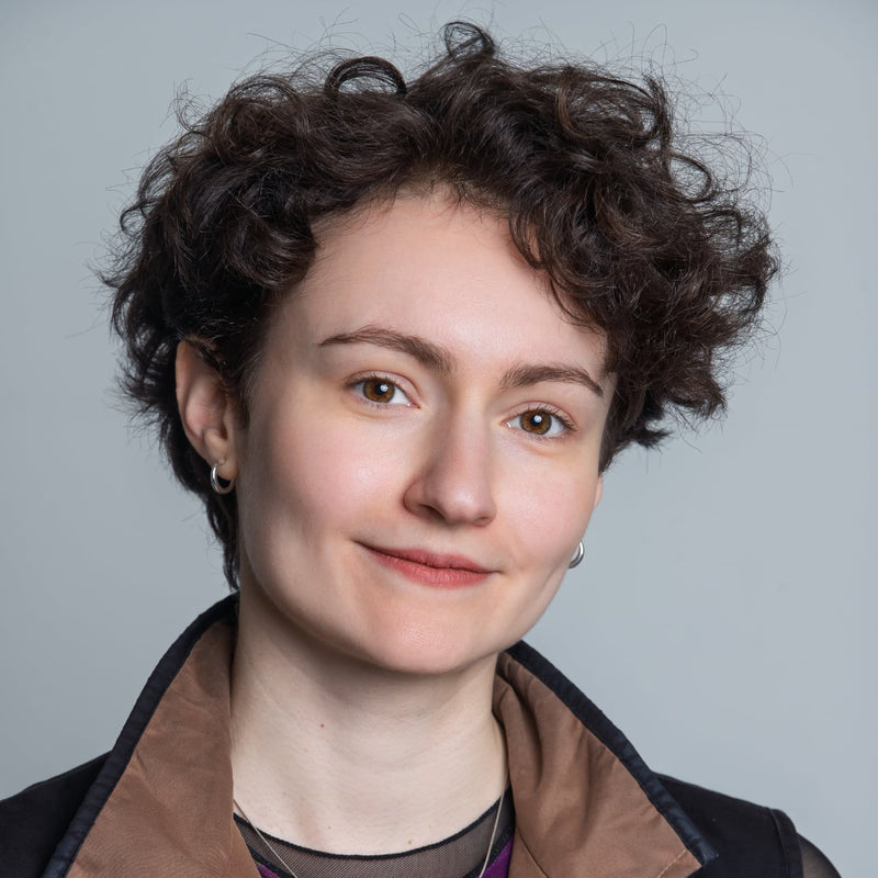 A close-up portrait of a woman with short curly brown hair, wearing a brown and black jacket, set against a solid light gray background.