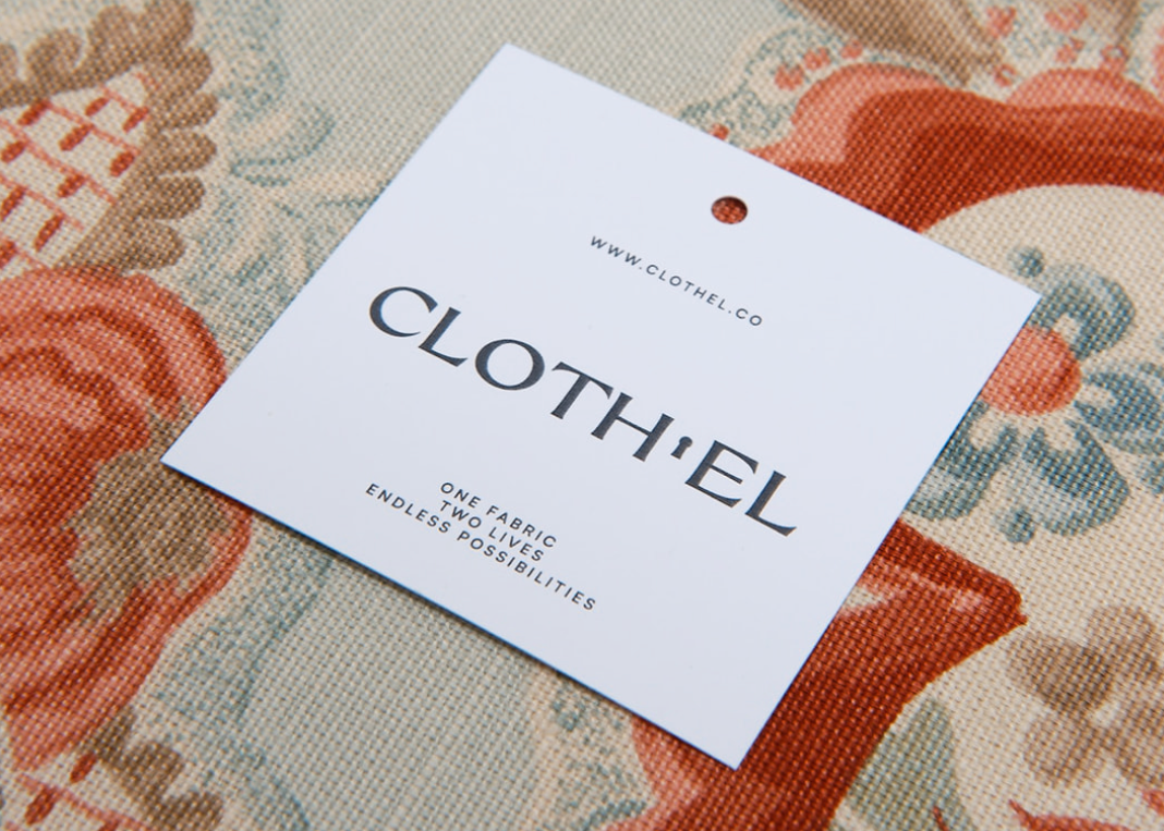A white paper hangtag with the brand name CLOTH'EL printed in dark gray, placed on a patterned fabric background with coral, beige, and muted blue floral motifs.