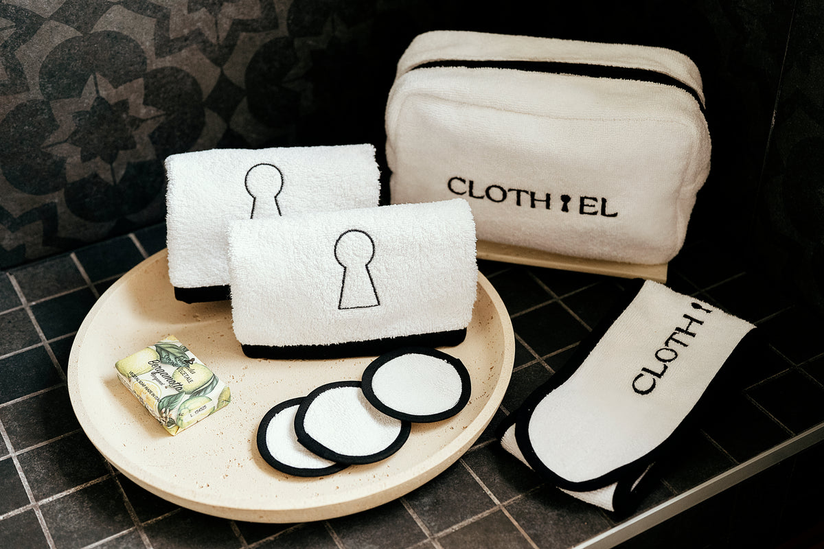 A set of white cloth wipes and towels with black trim, including a zippered pouch, a folded towel, a small towel, and a rectangular cloth, arranged on a beige tray with round wipe discs. The brand name CLOTHIEL is printed on the pouch and towel.