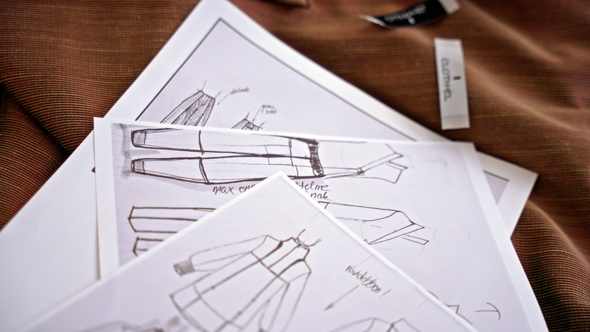 Several white sketch sheets with hand-drawn fashion illustrations are arranged on a brown textured fabric surface, with a small fabric tag visible nearby.