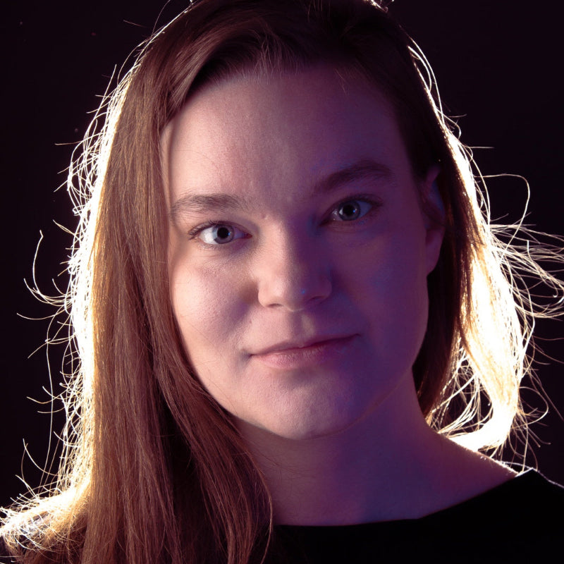 A close-up portrait of a young woman with long light brown hair, wearing a black top against a dark background with dramatic side lighting.